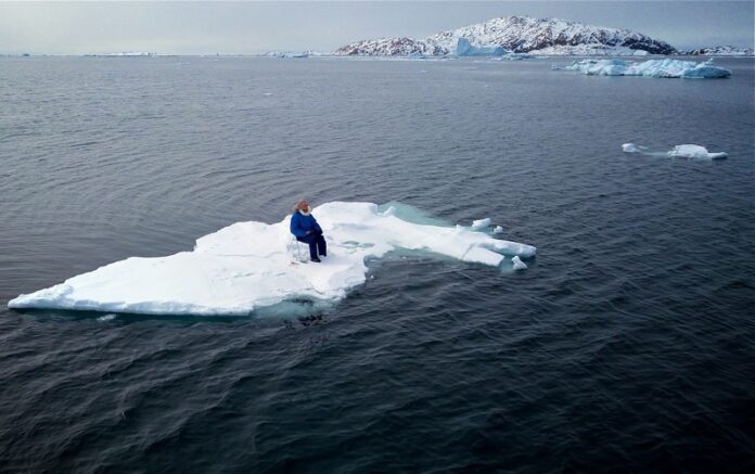 A man on a chair atop sea ice floating off west Greenland. The floe equates to the 15.69m² of Arctic sea ice lost due to his fossil fuel emissions flying between Australia and Greenland.
